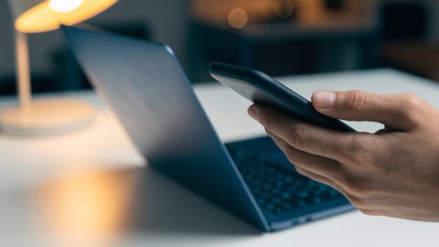 A hand holding a smartphone in front of a dark blue laptop on a white desk