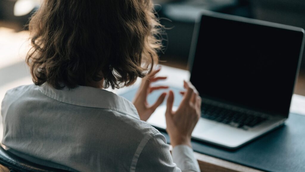A person seen from behind with hands raised at a silver laptop with a dark blank screen on a dark desk mat