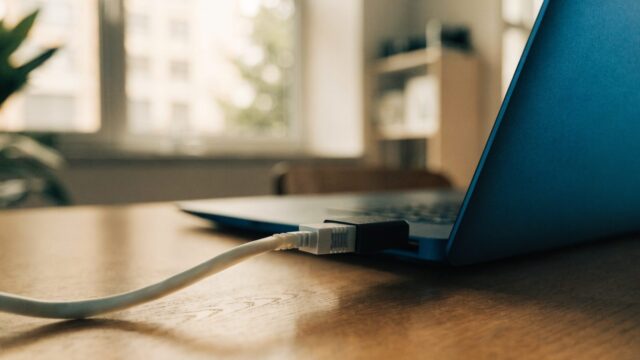 An ethernet cable being plugged into a USB-C adapter connected to a dark blue laptop on a wooden desk