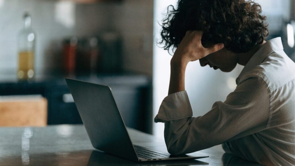 A person resting their forehead on one hand in front of a graphite black laptop on a kitchen table