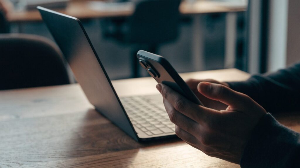 A hand holding a smartphone in front of a graphite black laptop on an office desk