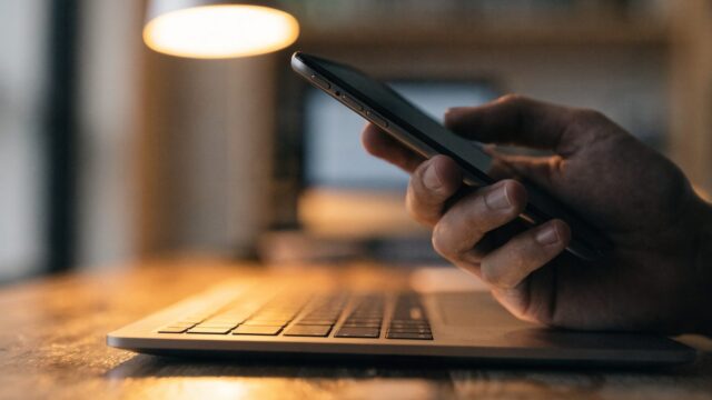 A hand holding a smartphone in front of a matte grey laptop on a wooden desk