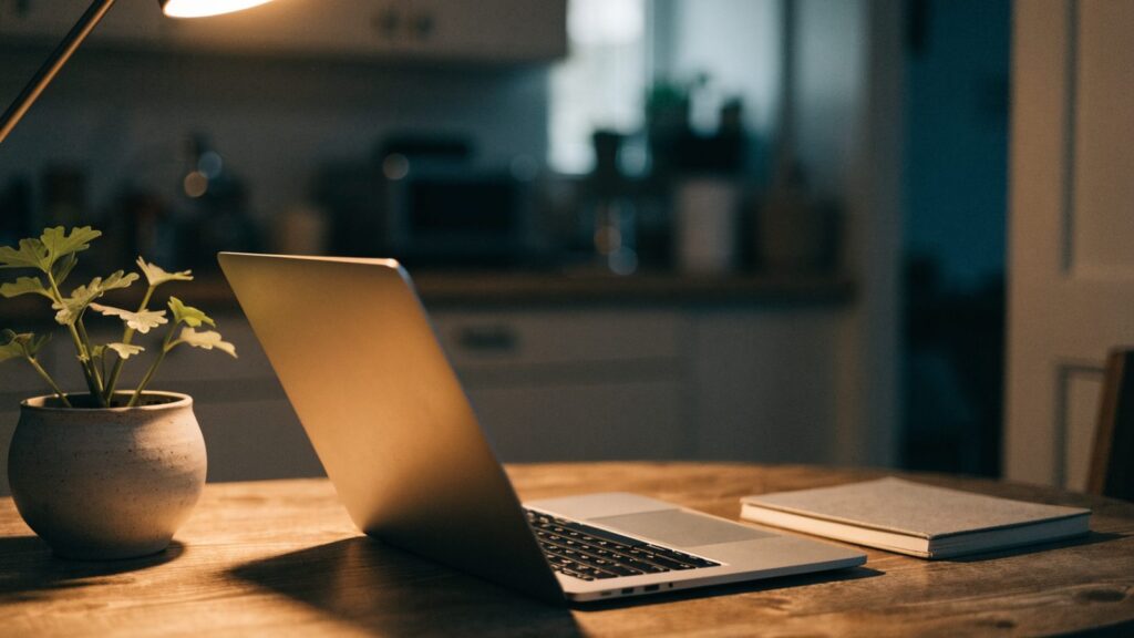 A silver laptop open on a kitchen table with a small plant and notebook beside it