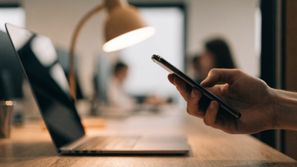 A hand holding a smartphone in front of a matte grey laptop on an office desk