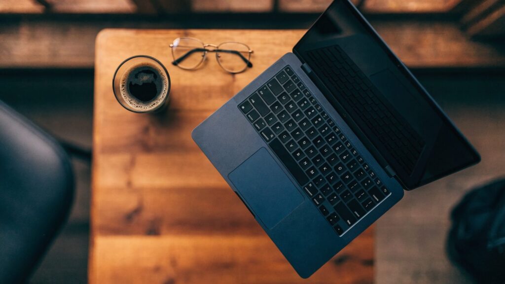 Top-down view of a dark blue laptop on a wooden desk with a coffee cup and glasses