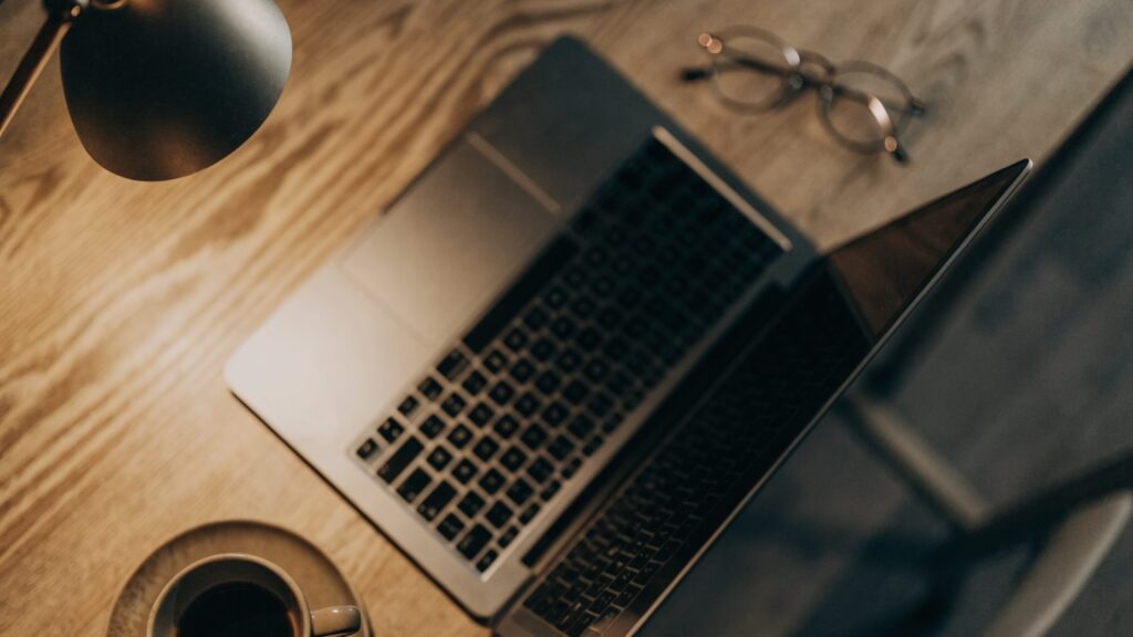 Top-down view of a silver laptop on a wooden desk with a coffee cup and glasses