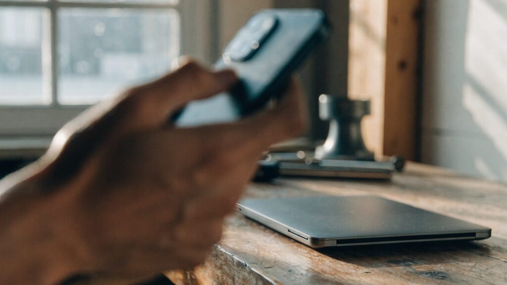 A hand holding a smartphone in front of a graphite black laptop on a workbench