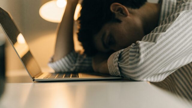 A person resting their forehead on one hand in front of a silver laptop on a white desk