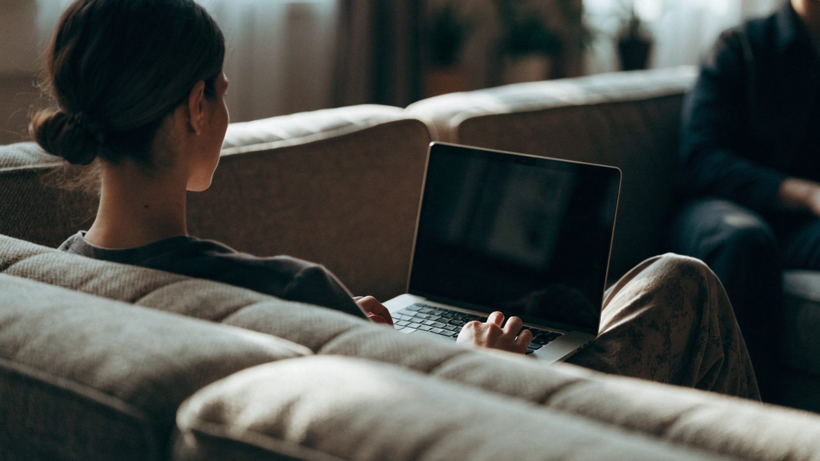A person seen from behind using a silver laptop on a sofa