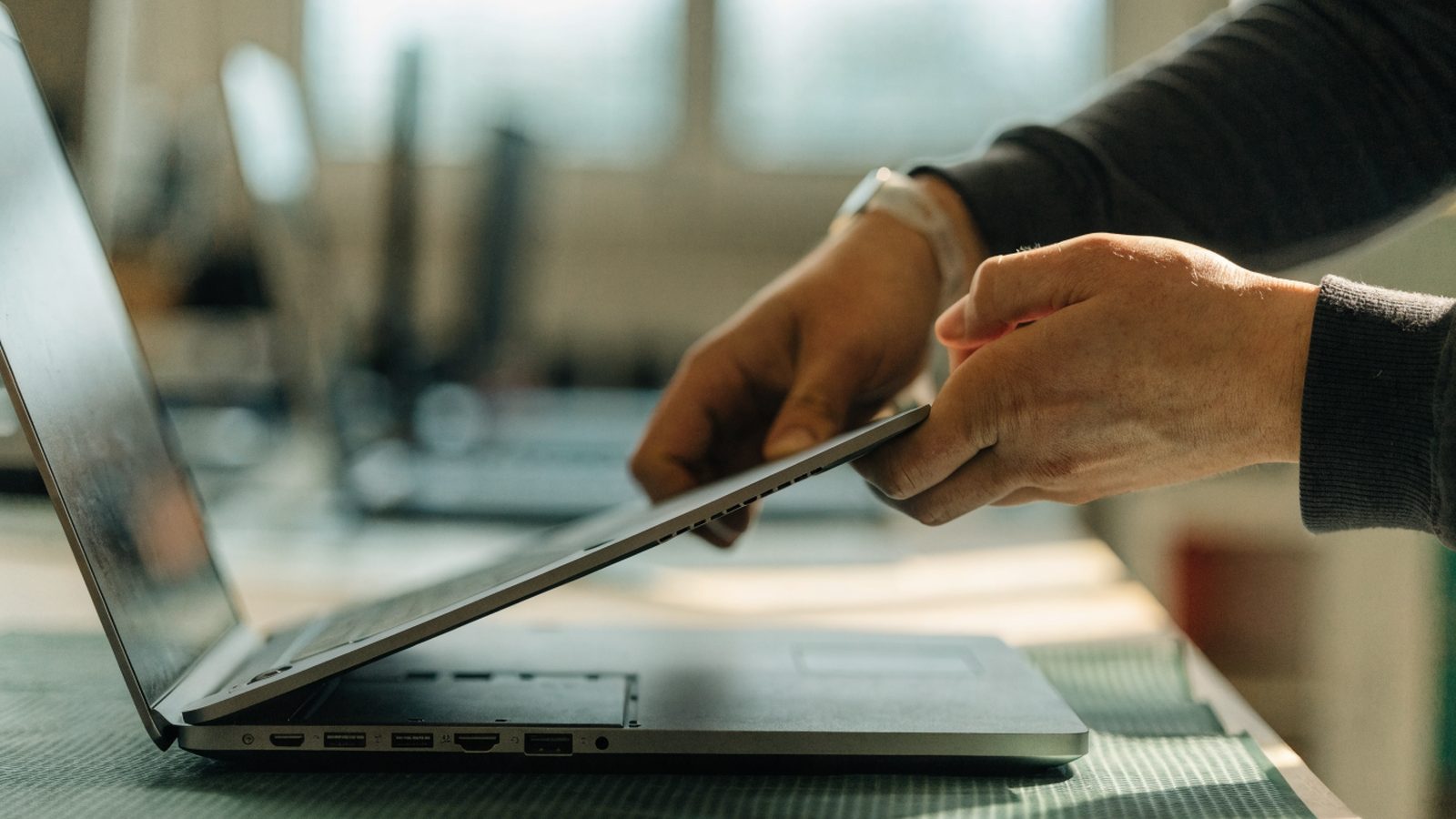 A technician lifting the bottom cover off a matte grey laptop on an anti-static mat