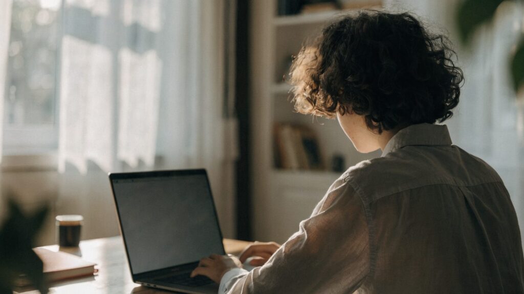 A person seen from behind working on a matte grey laptop in a bright home office