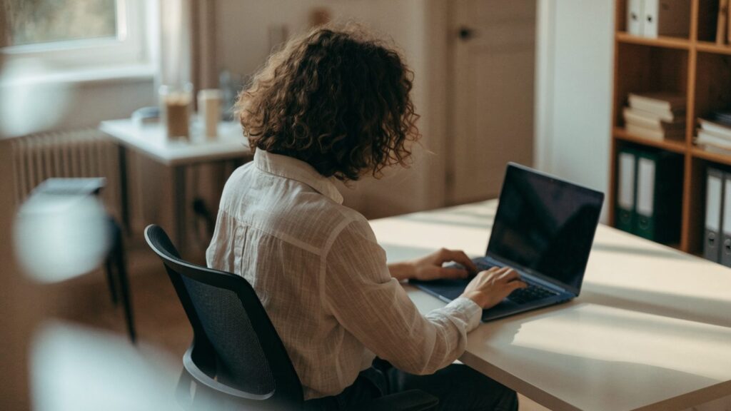 A person seen from behind working on a dark blue laptop in a bright home office