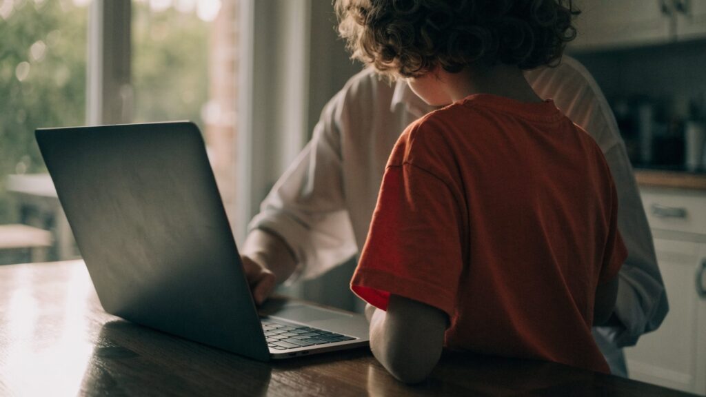 A parent and child seen from behind using a graphite black laptop together at a kitchen table