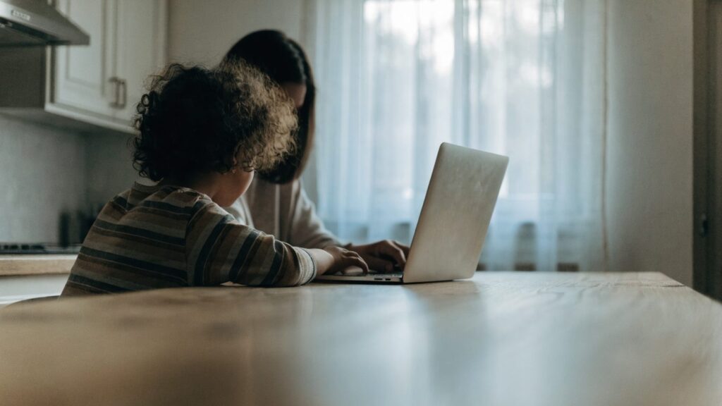 A parent and child seen from behind using a silver laptop together at a kitchen table