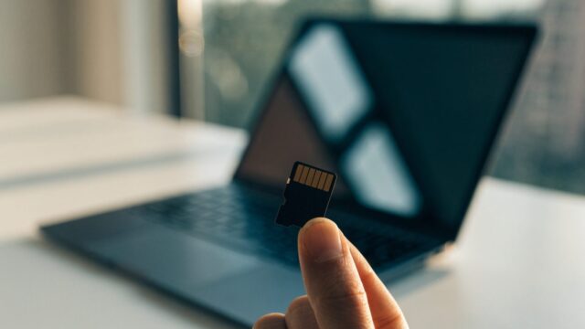 An SD card held between two fingers in front of a graphite black laptop on a white desk