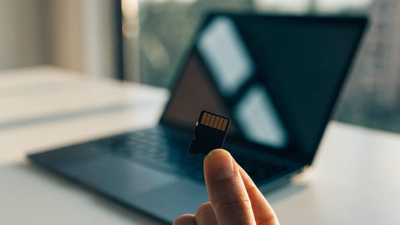 An SD card held between two fingers in front of a graphite black laptop on a white desk