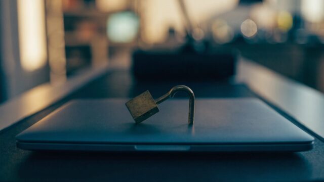 A small brass padlock resting on the closed lid of a dark blue laptop on a dark desk mat