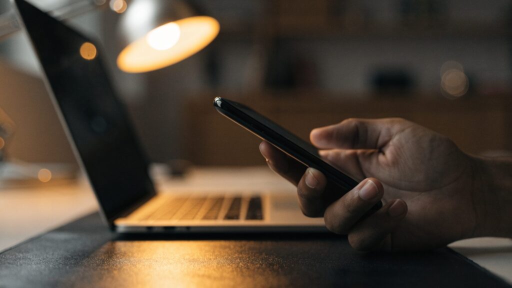 A hand holding a smartphone in front of a silver laptop on a dark desk mat