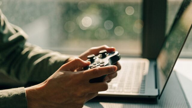 Hands holding a game controller in front of a matte grey laptop on a dark desk mat