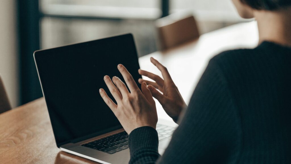 A person seen from behind with hands raised at a silver laptop with a dark blank screen on a wooden desk