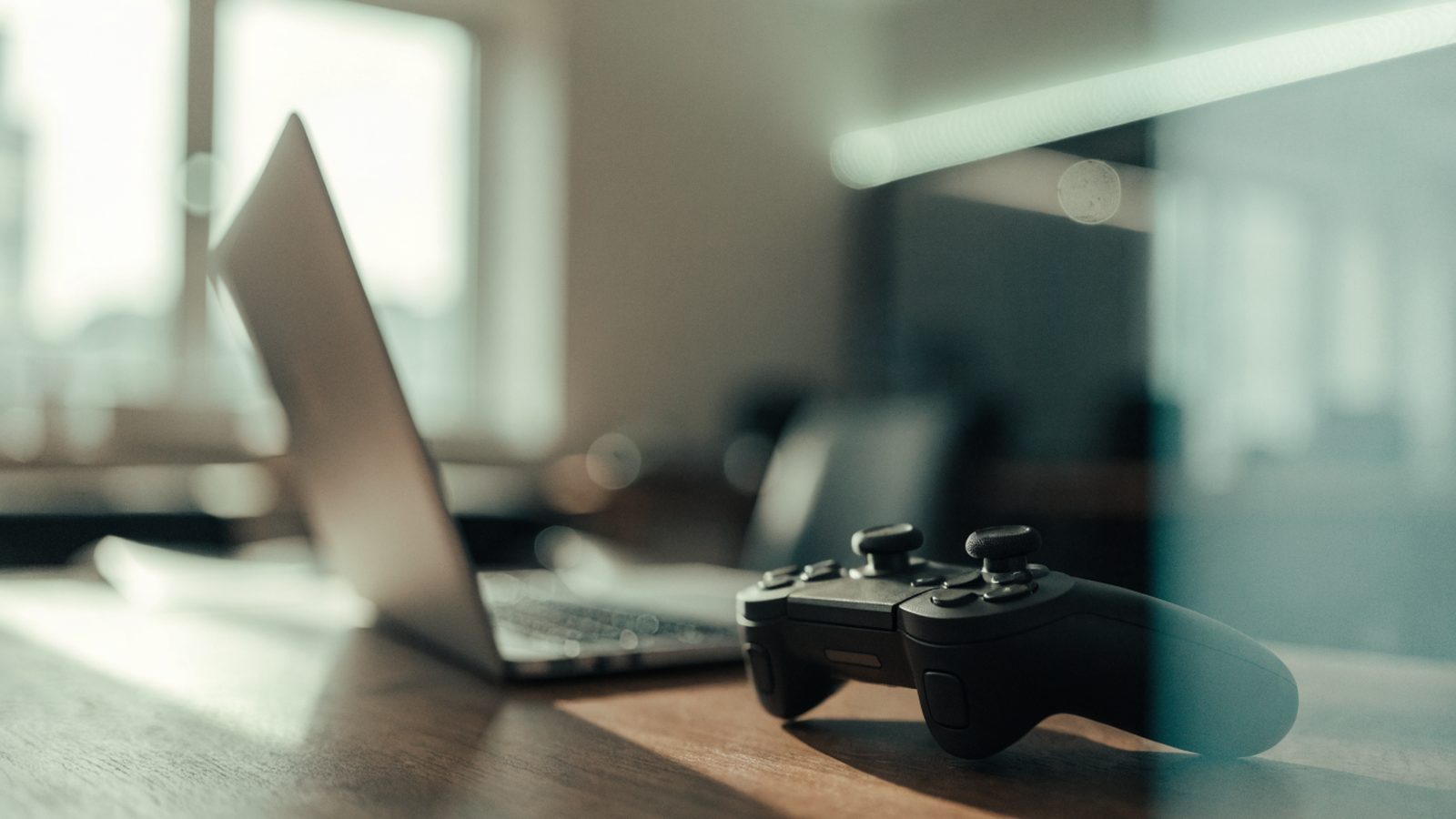 A game controller resting on a desk beside a silver laptop in a room with soft LED glow