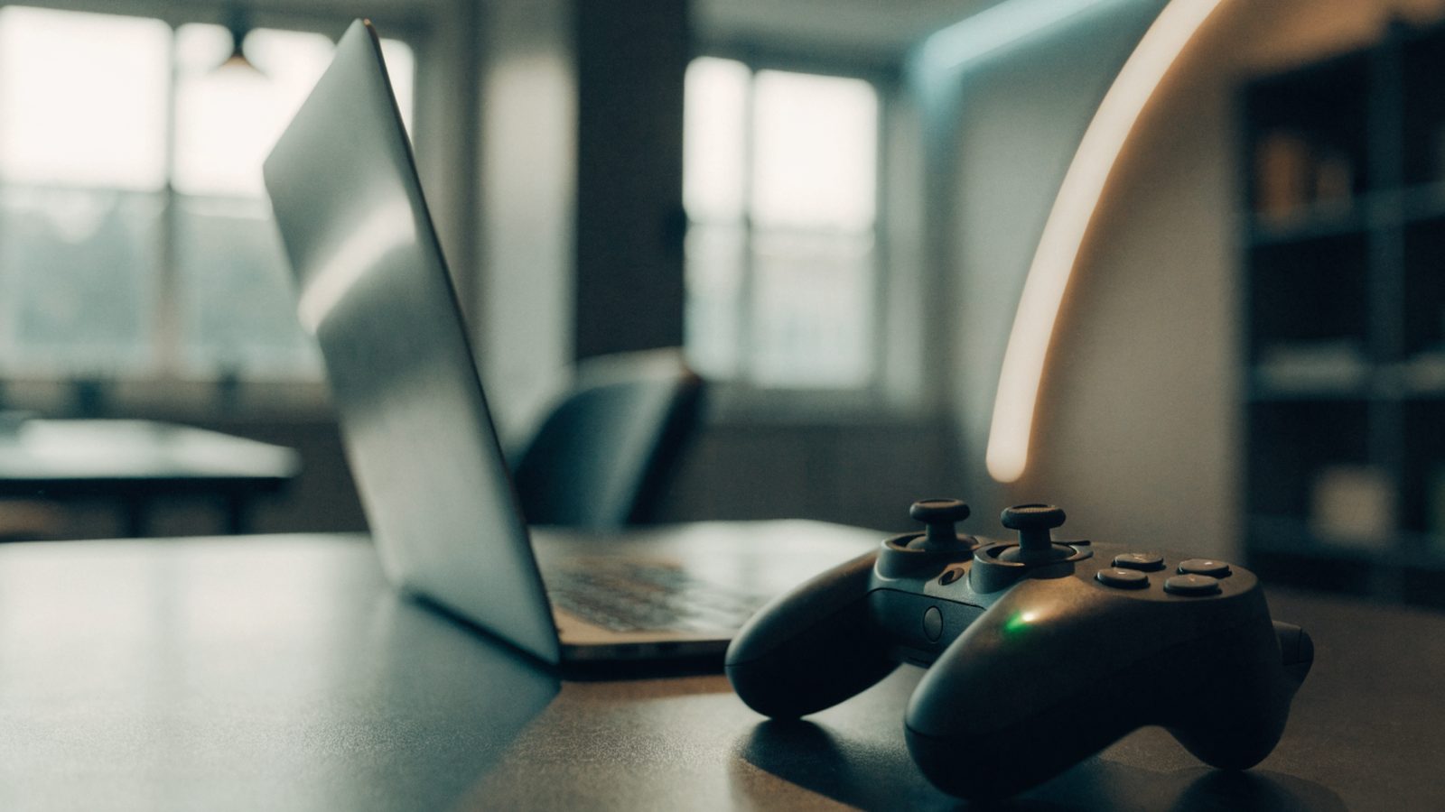 A game controller resting on a desk beside a silver laptop in a room with soft LED glow