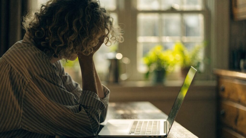 A person resting their forehead on one hand in front of a silver laptop on a kitchen table