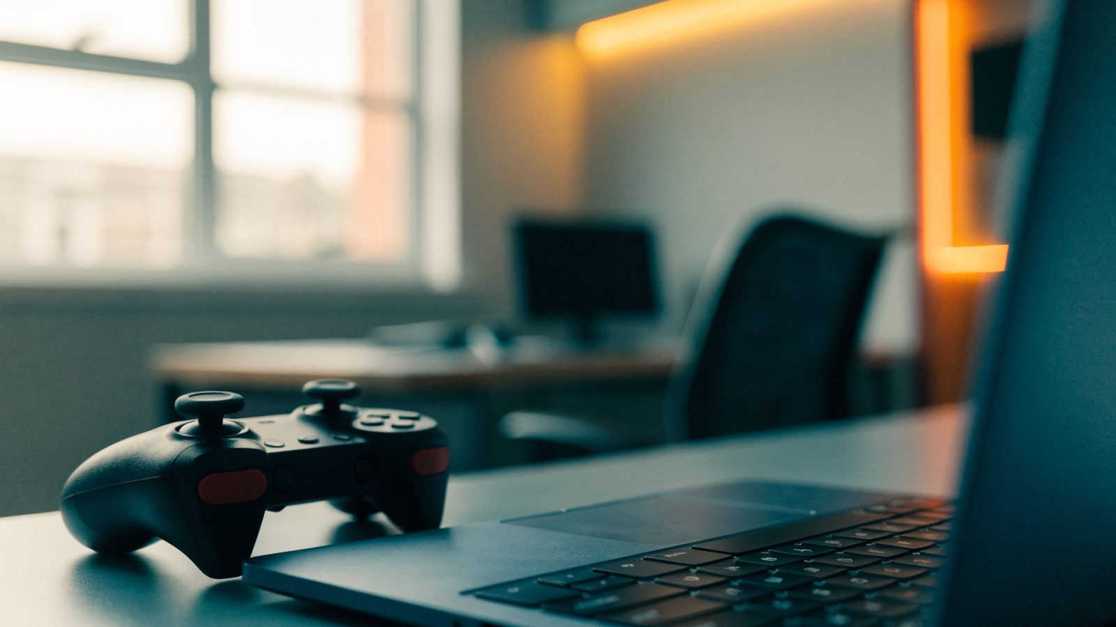 A game controller resting on a desk beside a dark blue laptop in a room with soft LED glow