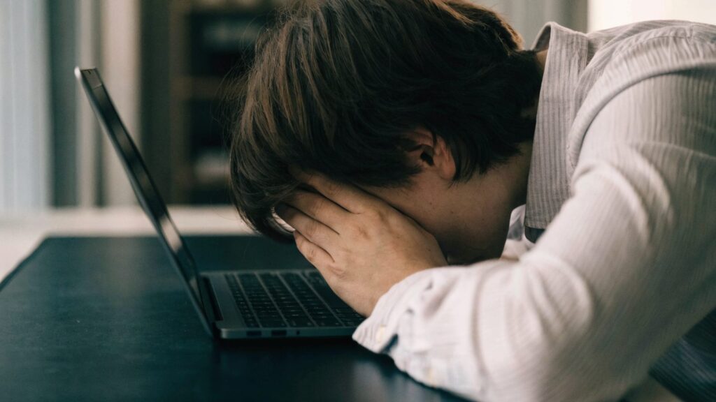 A person resting their forehead on one hand in front of a graphite black laptop on a dark desk mat