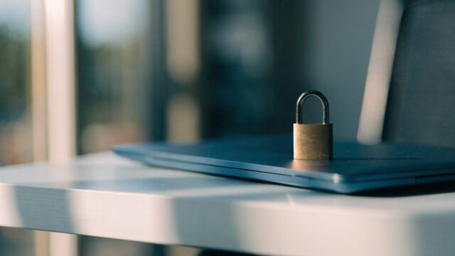 A small brass padlock resting on the closed lid of a dark blue laptop on a white desk