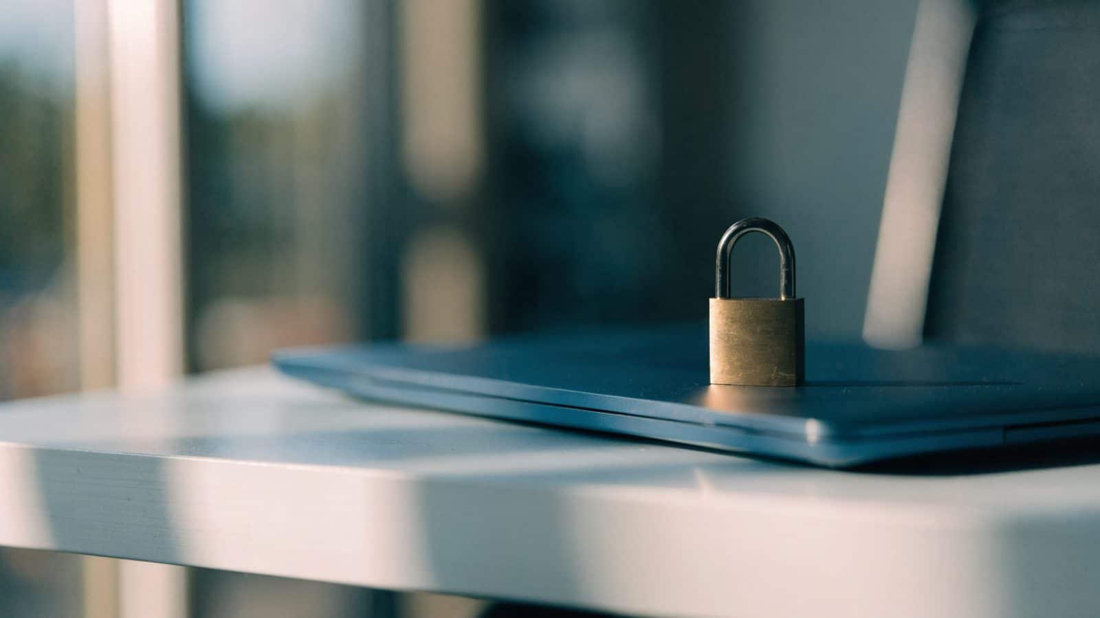 A small brass padlock resting on the closed lid of a dark blue laptop on a white desk