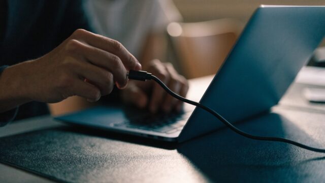 Hands unplugging a charging cable from a slim dark blue laptop on a dark desk mat