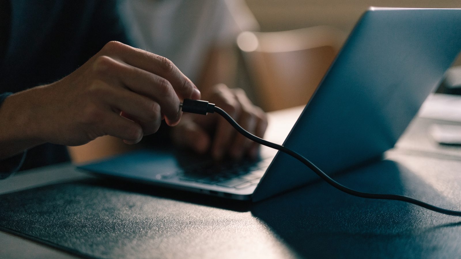 Hands unplugging a charging cable from a slim dark blue laptop on a dark desk mat