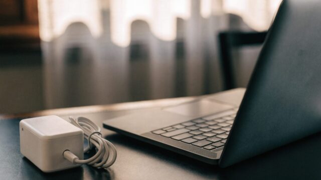 A graphite black laptop on a dark desk mat next to a compact white power adapter with its cable neatly coiled