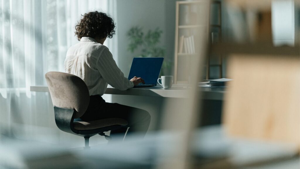 A person seen from behind working on a dark blue laptop in a bright home office