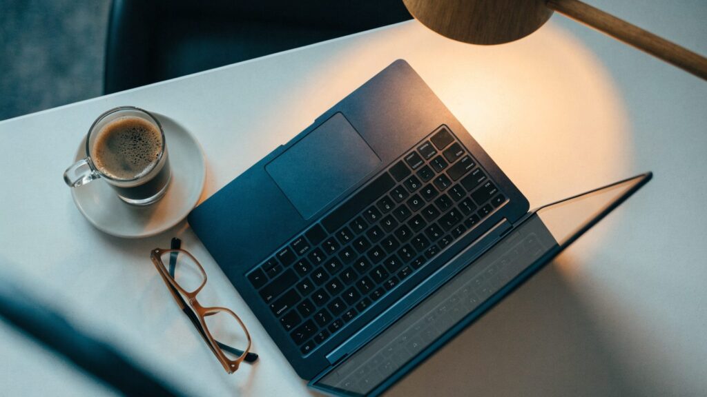 Top-down view of a dark blue laptop on a white desk with a coffee cup and glasses