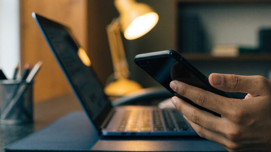 A hand holding a smartphone in front of a dark blue laptop on a dark desk mat