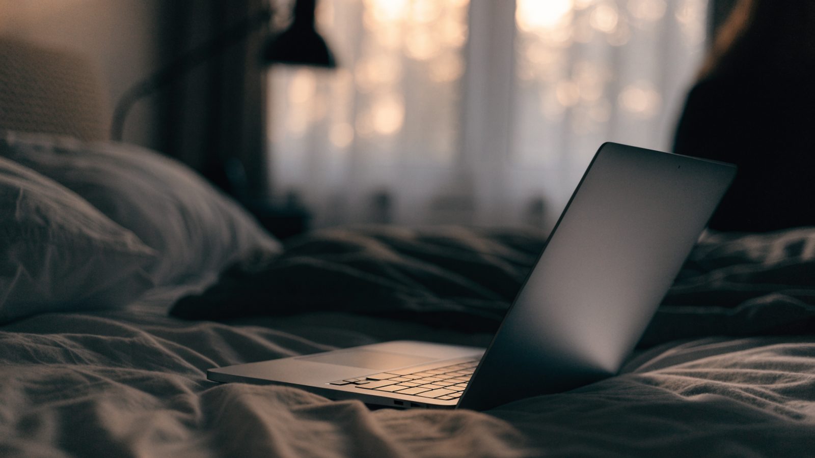 A matte grey laptop glowing softly on a bed in a dark room