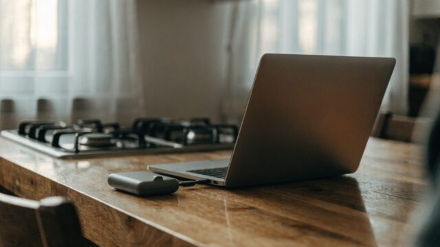A small external hard drive connected by cable to a matte grey laptop on a kitchen table