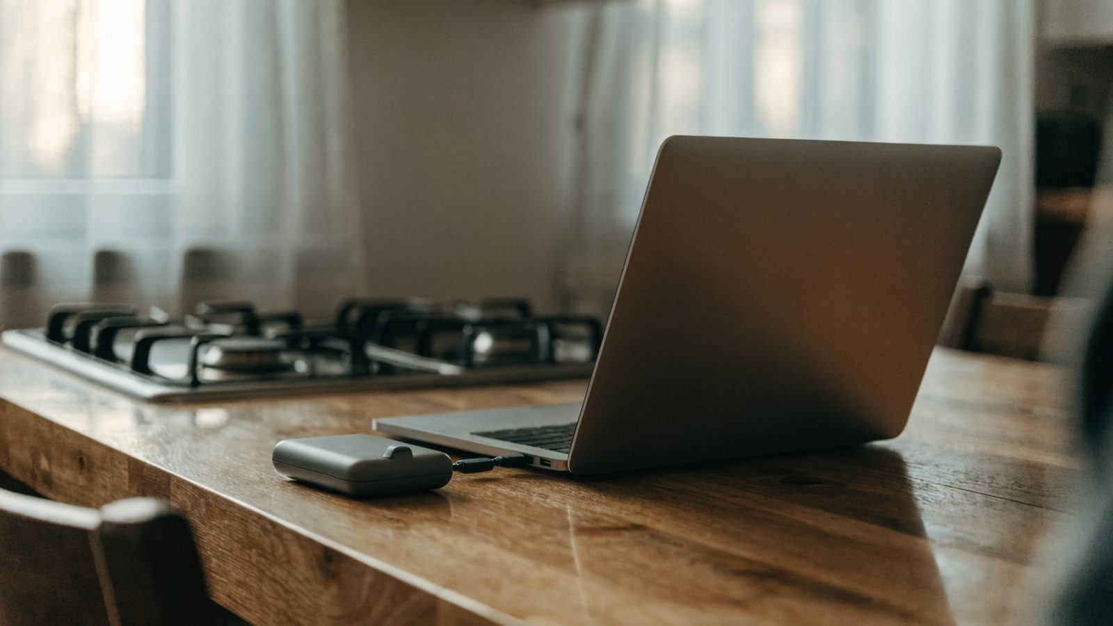 A small external hard drive connected by cable to a matte grey laptop on a kitchen table