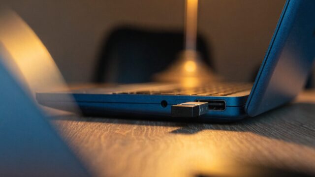 A USB flash drive plugged into the side port of a dark blue laptop on a wooden desk