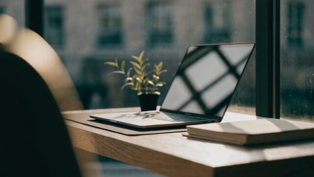 A matte grey laptop open on a dark desk mat with a small plant and notebook beside it