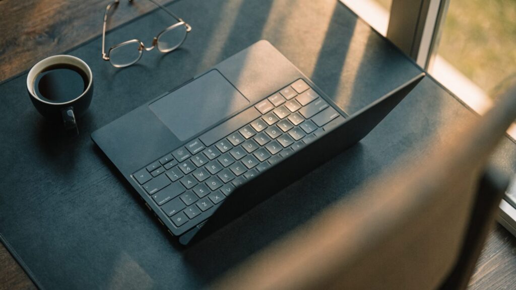Top-down view of a graphite black laptop on a dark desk mat with a coffee cup and glasses