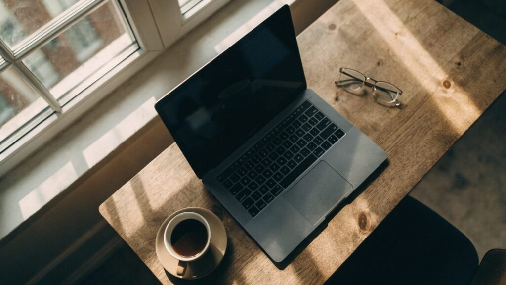 Top-down view of a matte grey laptop on a wooden desk with a coffee cup and glasses