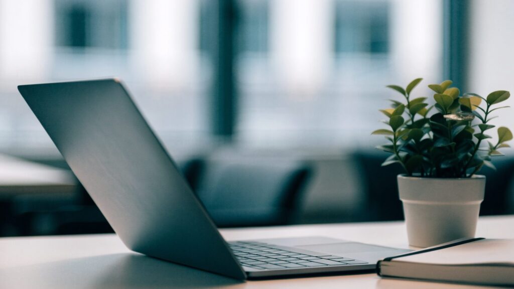 A matte grey laptop open on an office desk with a small plant and notebook beside it