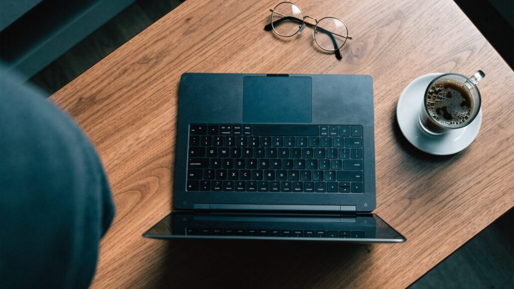 Top-down view of a graphite black laptop on a wooden desk with a coffee cup and glasses