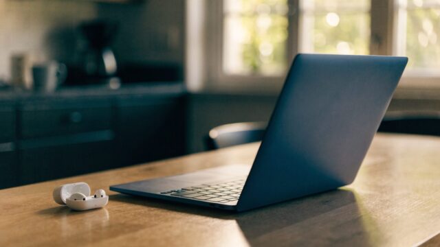 White wireless earbuds in their open case beside a dark blue laptop on a kitchen table