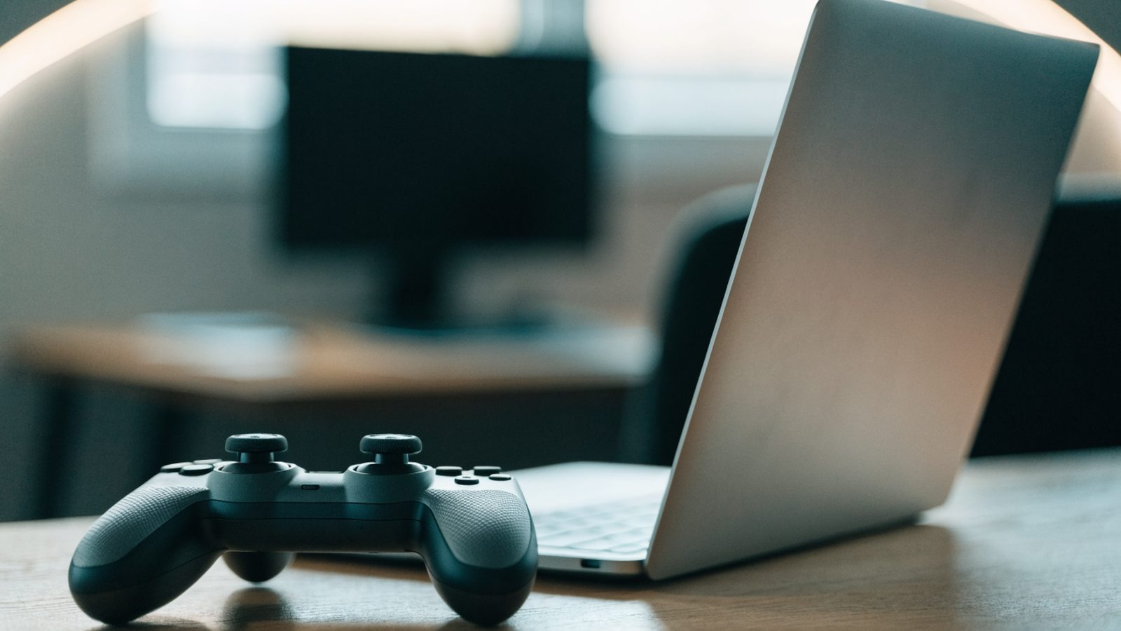 A game controller resting on a desk beside a silver laptop in a room with soft LED glow