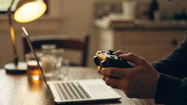 Hands holding a game controller in front of a silver laptop on a kitchen table