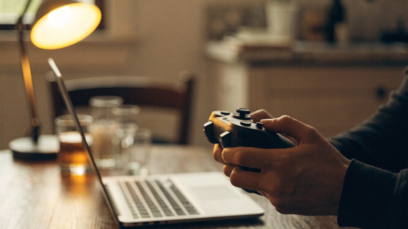 Hands holding a game controller in front of a silver laptop on a kitchen table